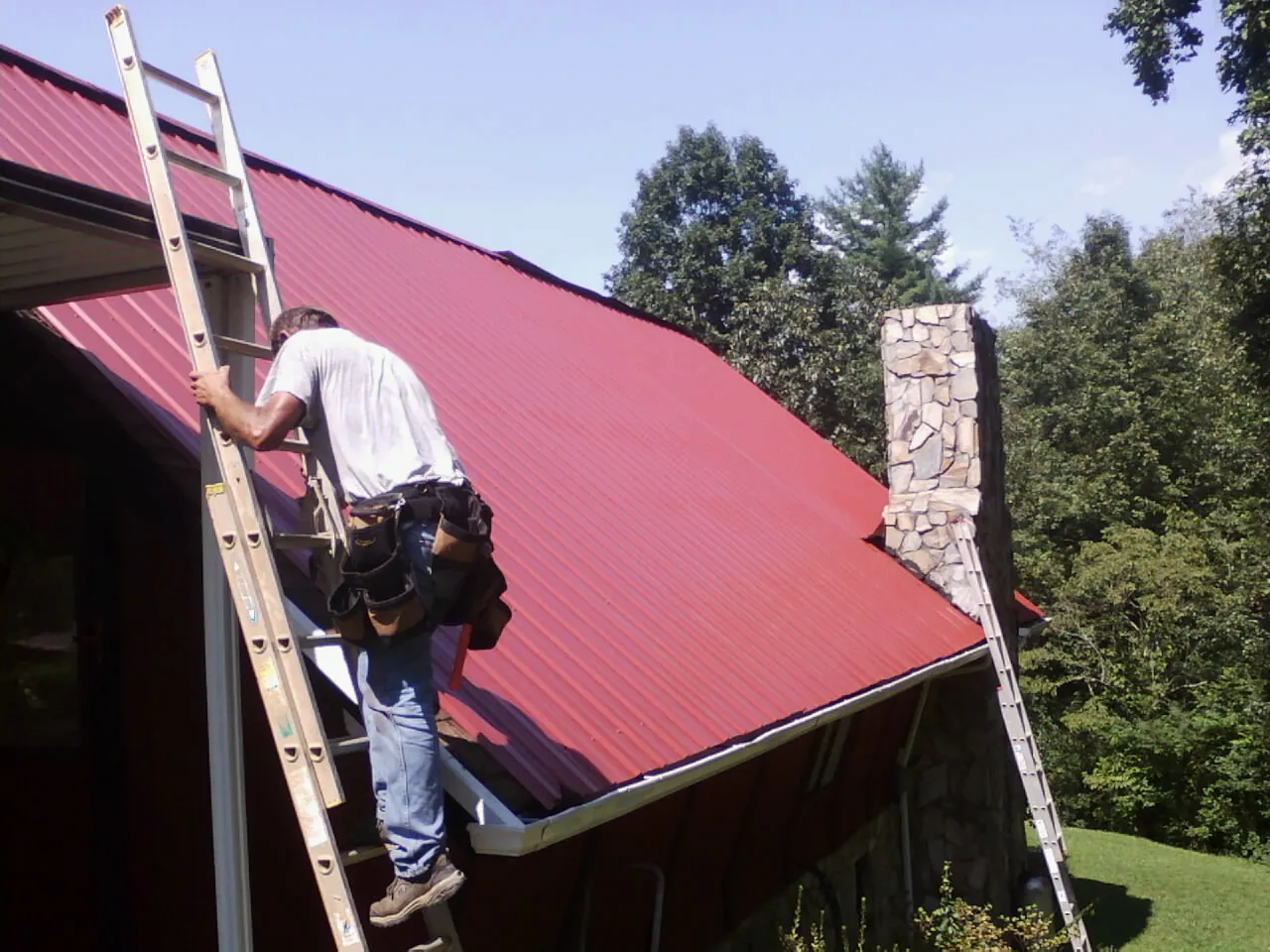 Roofer on a ladder beside a red metal roof and stone chimney — Soffit & Fascia Repair by Roofing Brothers in South Vienna